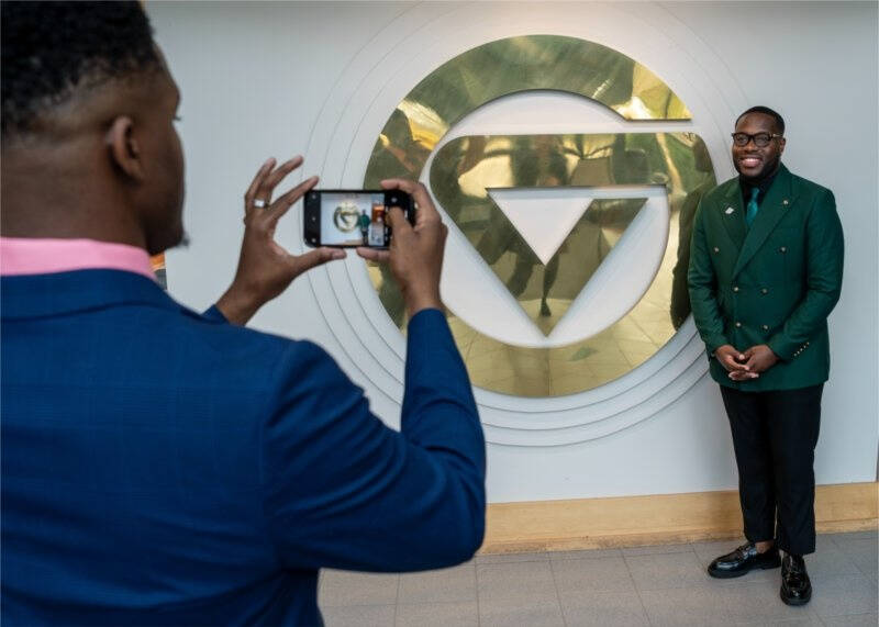 A man in a blue suit is taking a photo with his smartphone of another man who is standing in front of a large, gold and white circular GVSU logo on a wall. The man being photographed is wearing a green double-breasted blazer and black pants.